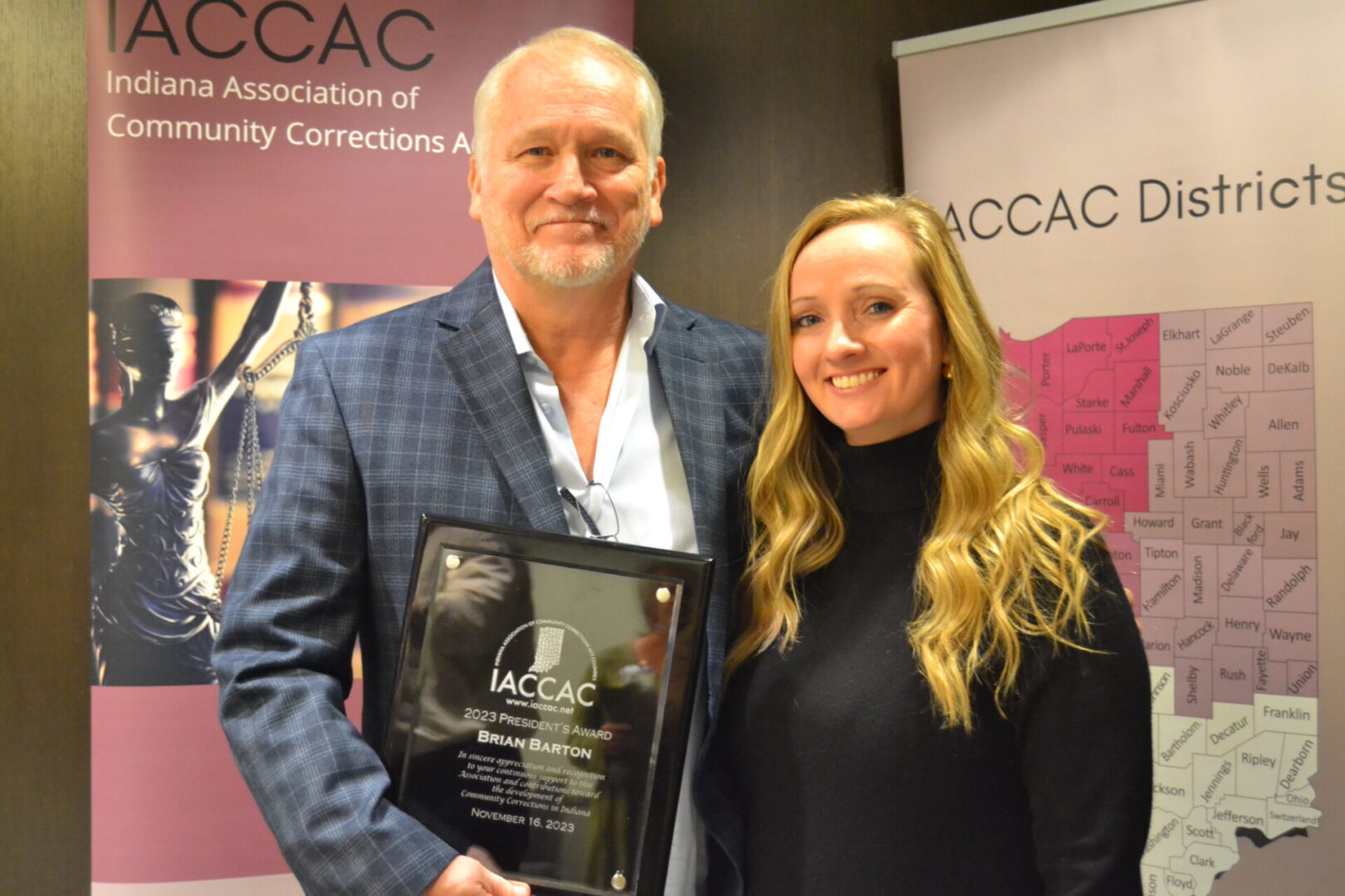 A man and woman posing for a photo with an award in front of a banner.